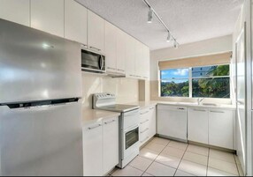 Una cocina moderna con armarios blancos, un refrigerador de acero inoxidable, microondas, estufa y lavavajillas. Hay una ventana que ofrece vistas a zonas verdes.