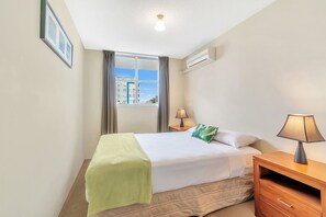A hotel room with a bed, bedside table, lamp, and a window with curtains. - Tradewinds Apartments (Coffs Harbour)