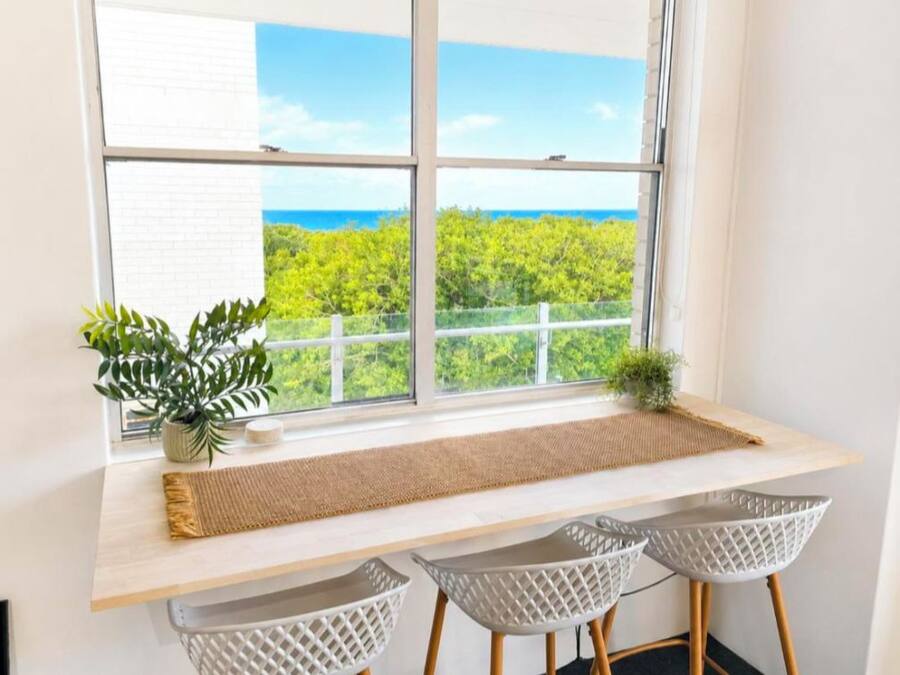 Quarto com uma grande janela que dá para uma paisagem verde e o mar. Há uma escrivaninha de madeira com um tapete tecido, duas cadeiras brancas e um pequeno vaso de planta.