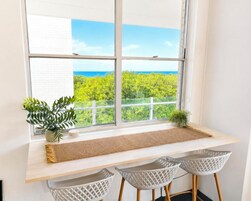A room with a large window overlooking a green landscape and the sea. There is a wooden desk with a woven mat, two white chairs, and a small potted plant.