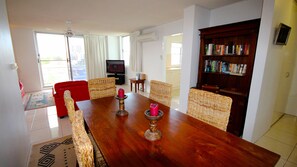 A dining area with a wooden table, wicker chairs, a red sofa, and a bookshelf. - Tradewinds Apartments (Coffs Harbour)