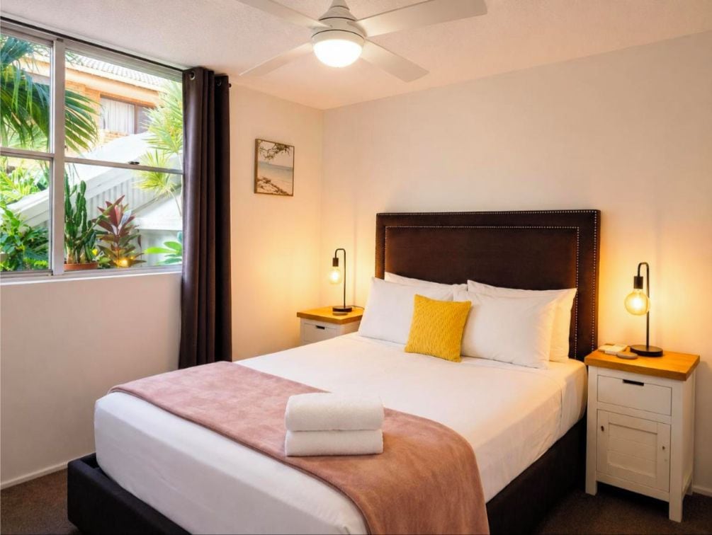 Quarto de hotel com cama, mesas de cabeceira, ventilador de teto e vista para o verde lá fora.