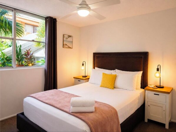 A hotel room with a bed, bedside tables, a ceiling fan, and a view of greenery outside.