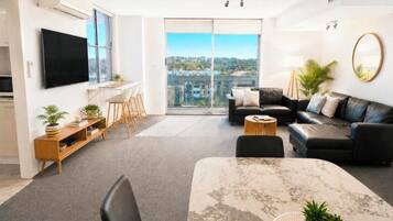 A modern living room with a flat-screen TV, a dining table with chairs, a sofa, and a large window offering a city view.
