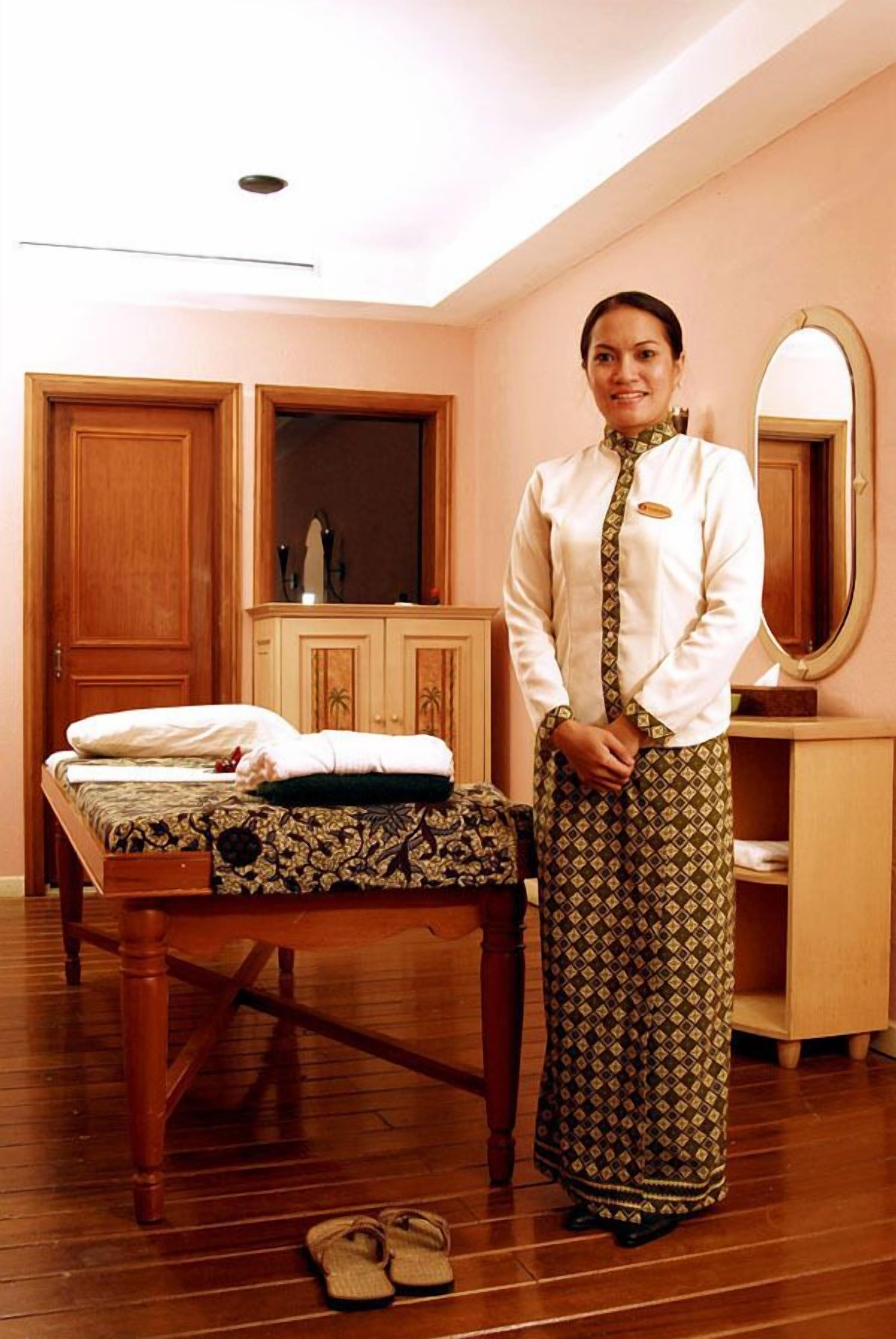 A room with a wooden bed, a wooden dresser, a mirror, and a person standing by the bed.