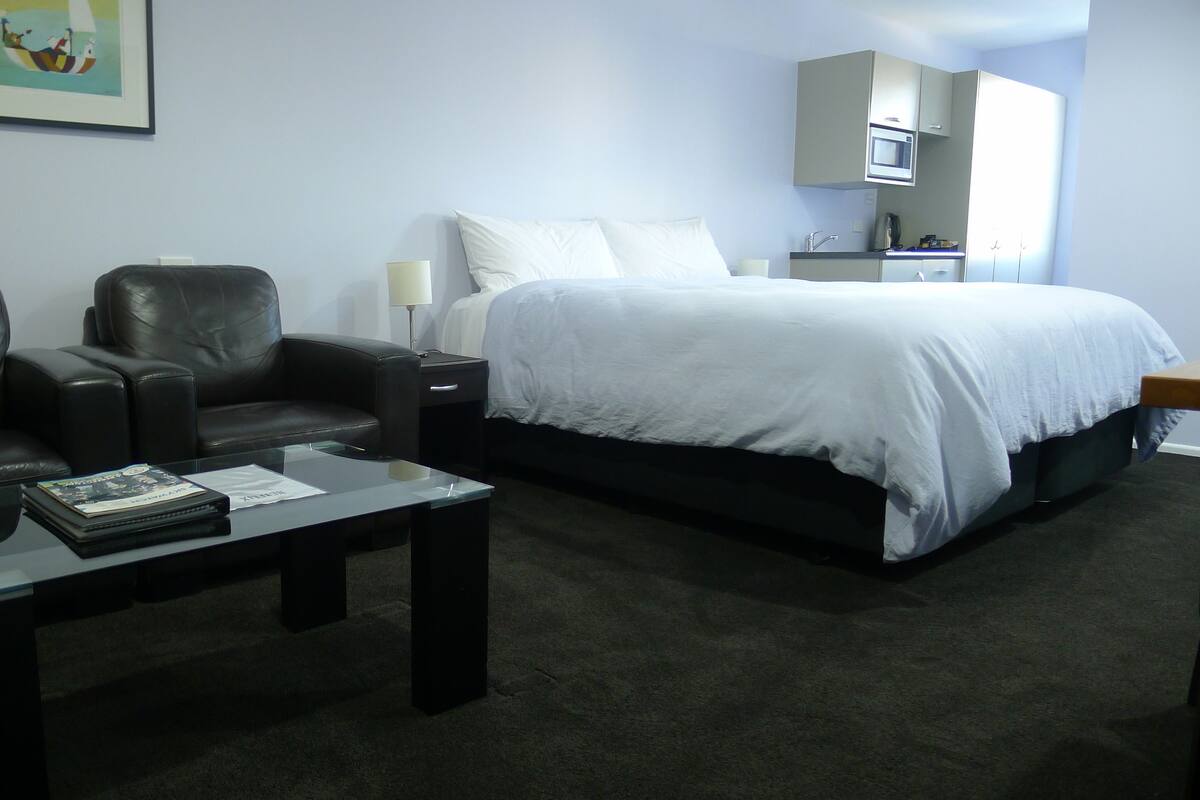 A hotel room with a bed, two leather chairs, a small table with a book, a nightstand, and a microwave above the bed.