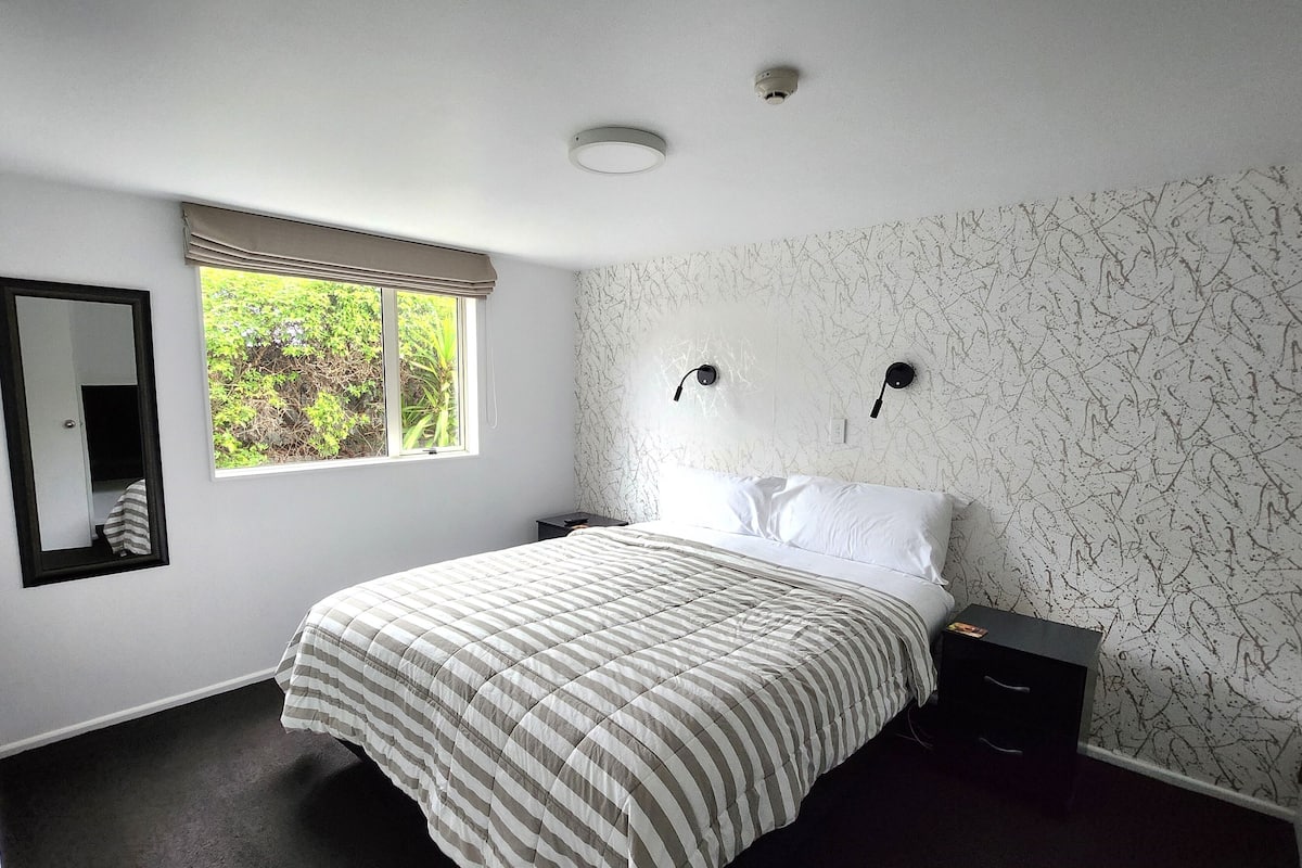 A bedroom with a bed, a nightstand, a mirror, and a window with a view of greenery.