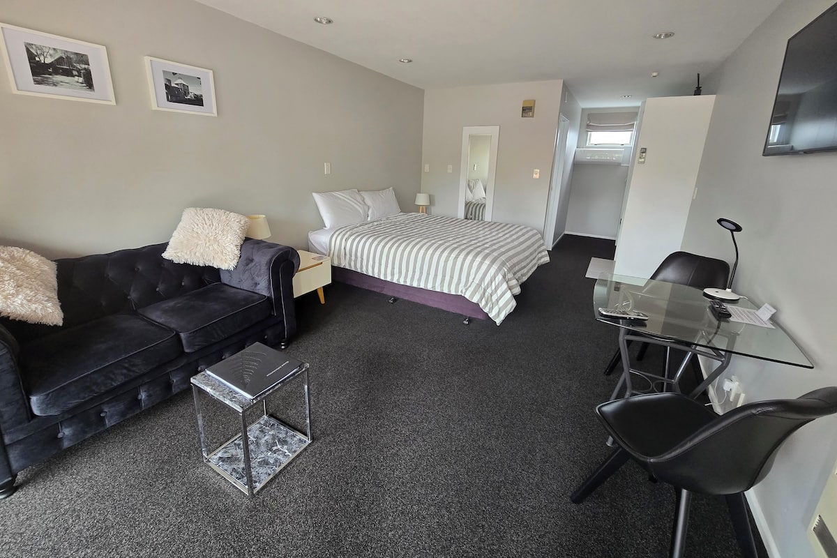 A modern hotel room with a black leather sofa, a bed with striped bedding, a glass desk with a chair, and a small table.