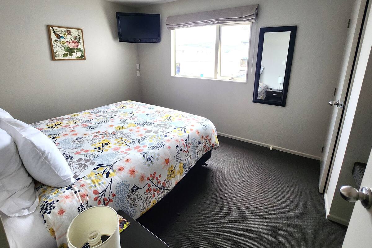 A bedroom with a bed, a flat-screen TV, a window with curtains, a mirror, and a door.