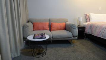 A hotel room with a grey sofa, two orange pillows, a small round table with a magazine, a black metal chair, a white lamp, and a bed with a striped blanket.