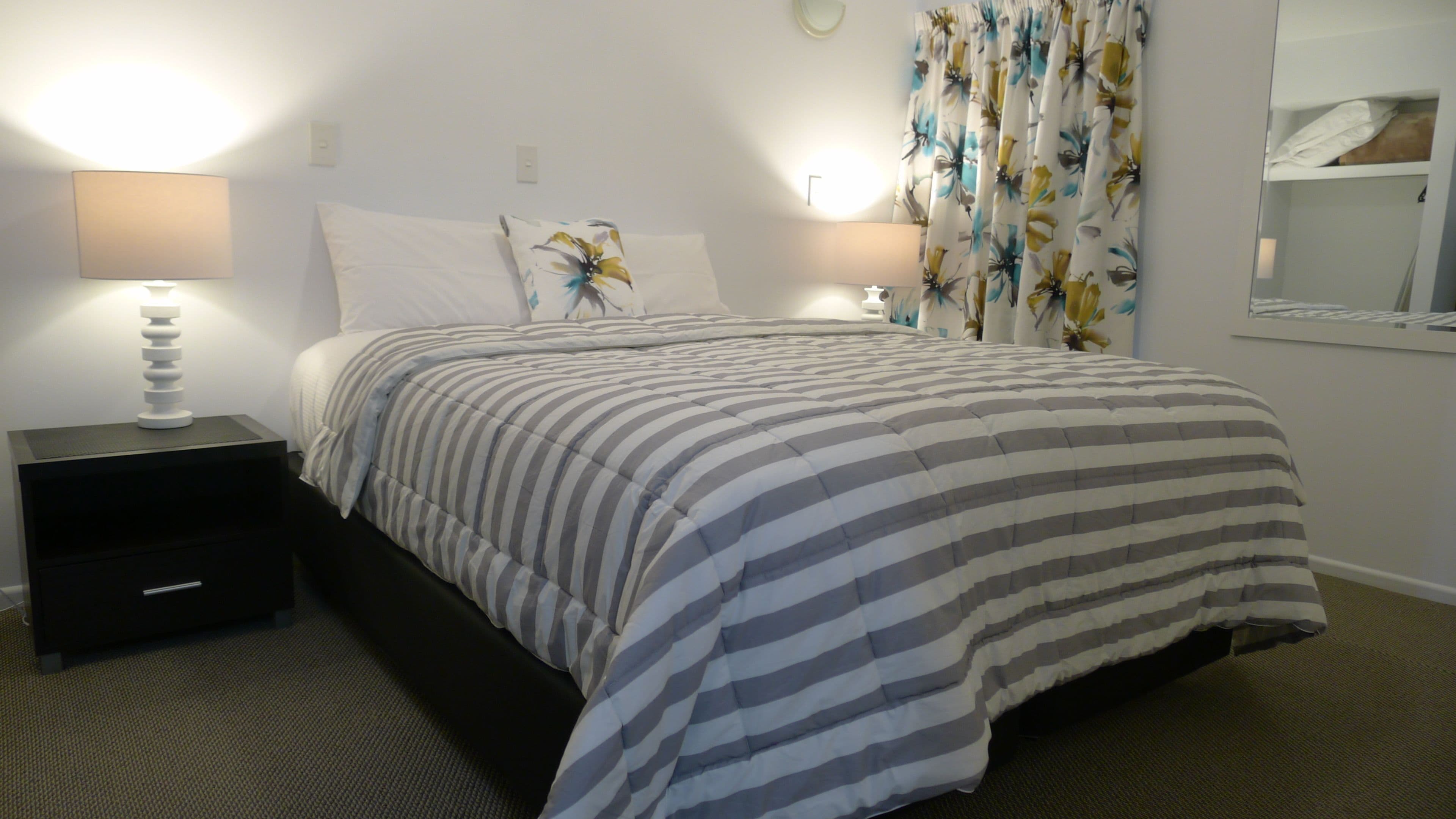A hotel room with a bed, two bedside lamps, a nightstand, a mirror, and a floral curtain.