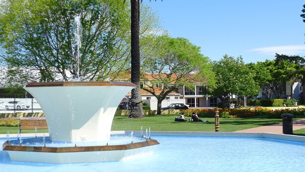 Una fuente con una columna central y chorros de agua, rodeada por una piscina. Al fondo, se ven árboles, un césped y edificios.