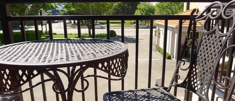 A balcony with a round metal table and a chair, overlooking a parking lot and buildings.