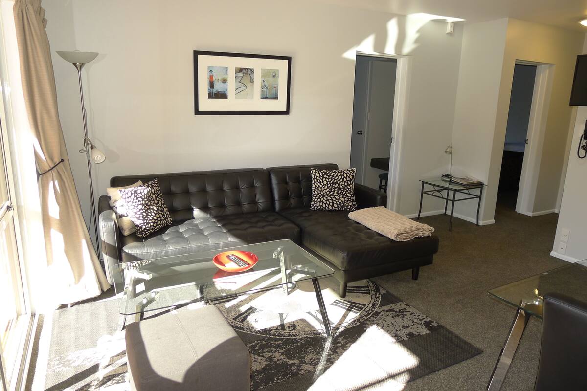 A living room with a black leather sofa, a glass coffee table, and a patterned rug. There is a framed artwork on the wall and a door leading to another room.