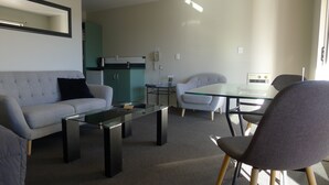 Una sala moderna con un sofá, una mesita de centro de vidrio, una mesa de comedor con sillas, una kitchenette y un pequeño espacio de oficina.