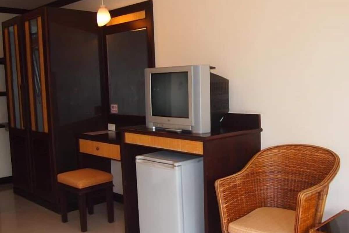A compact room with a wooden desk, a television, a small refrigerator, a wicker chair, and a hanging light fixture.