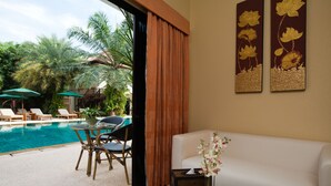 Une chambre d’hôtel avec vue sur une piscine, un canapé blanc, une table d’appoint en bois ornée d’un vase de fleurs et deux tableaux floraux encadrés accrochés au mur.