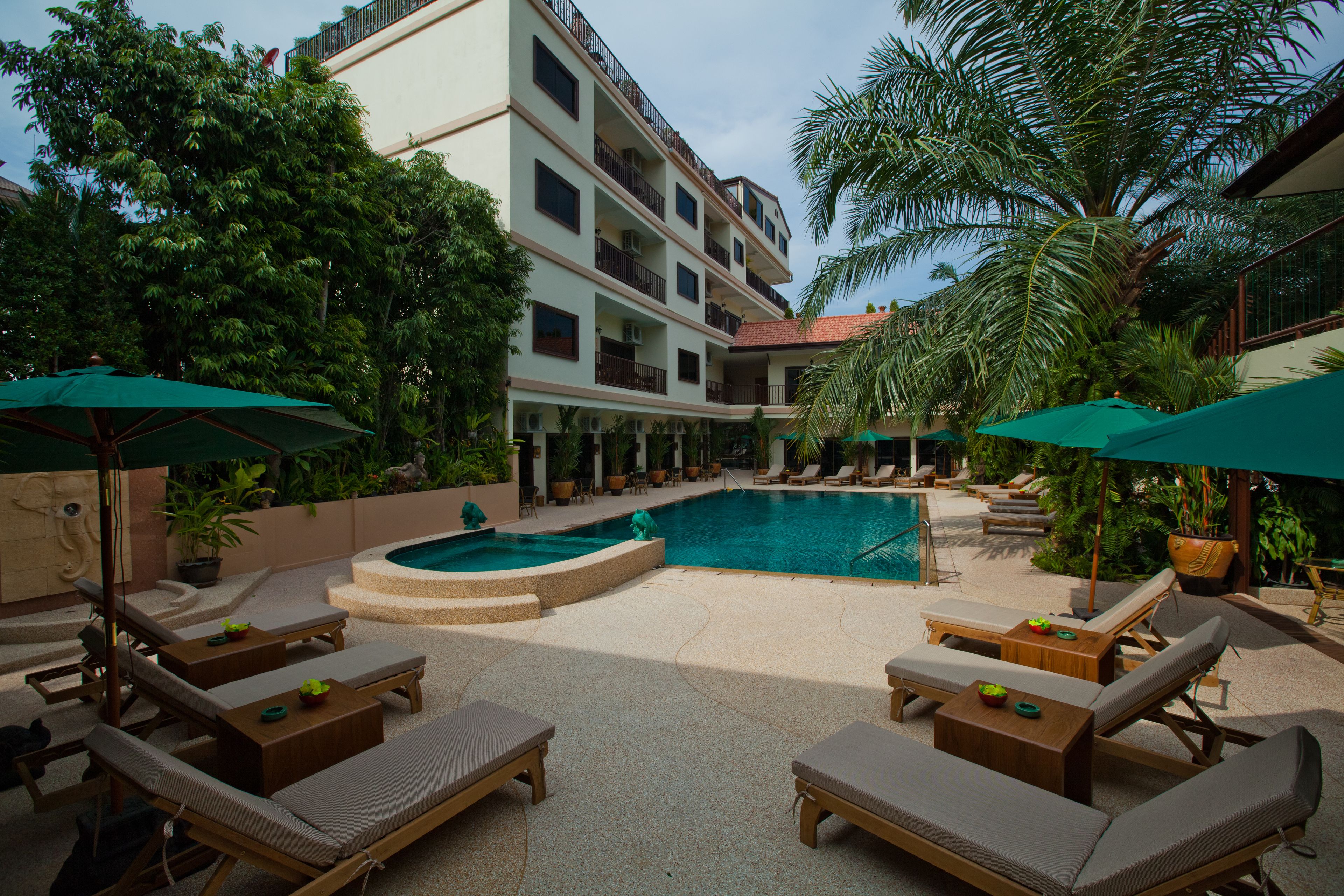 A pool area with lounge chairs and umbrellas, surrounded by greenery and a multi-story building.