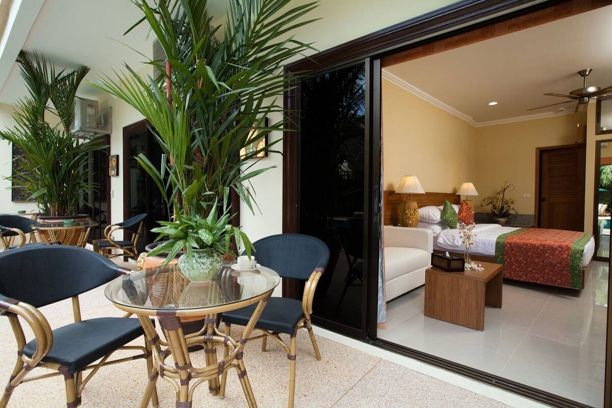 Ein modernes Hotelzimmer mit Glastür zu einem Balkon, einem Bett mit roter und grüner Bettdecke, einem kleinen Nachttisch aus Holz und einer Topfpflanze.