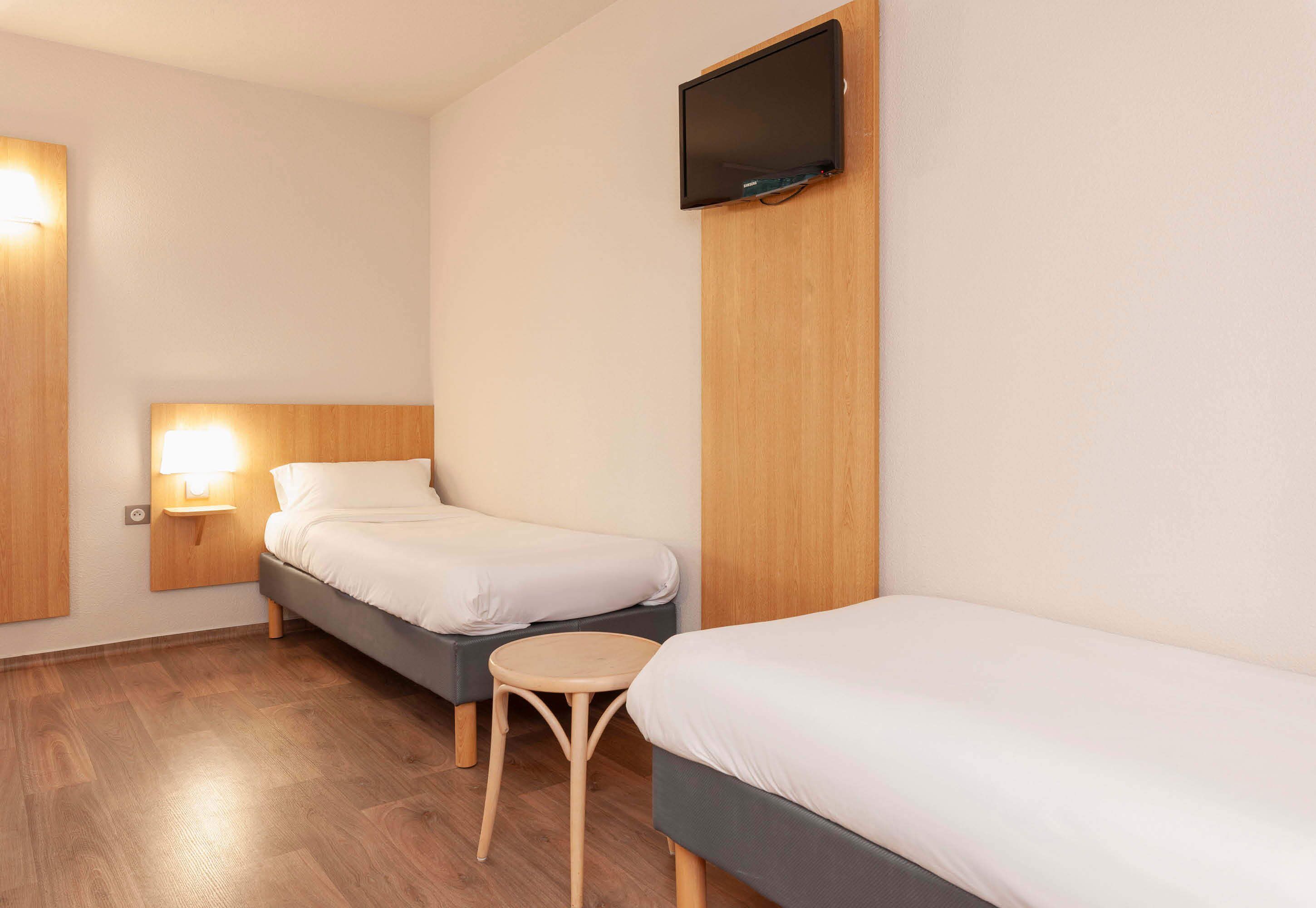 Una habitación de hotel con dos camas individuales, una mesita redonda pequeña, un televisor montado en la pared y una mesita de noche con lámpara.