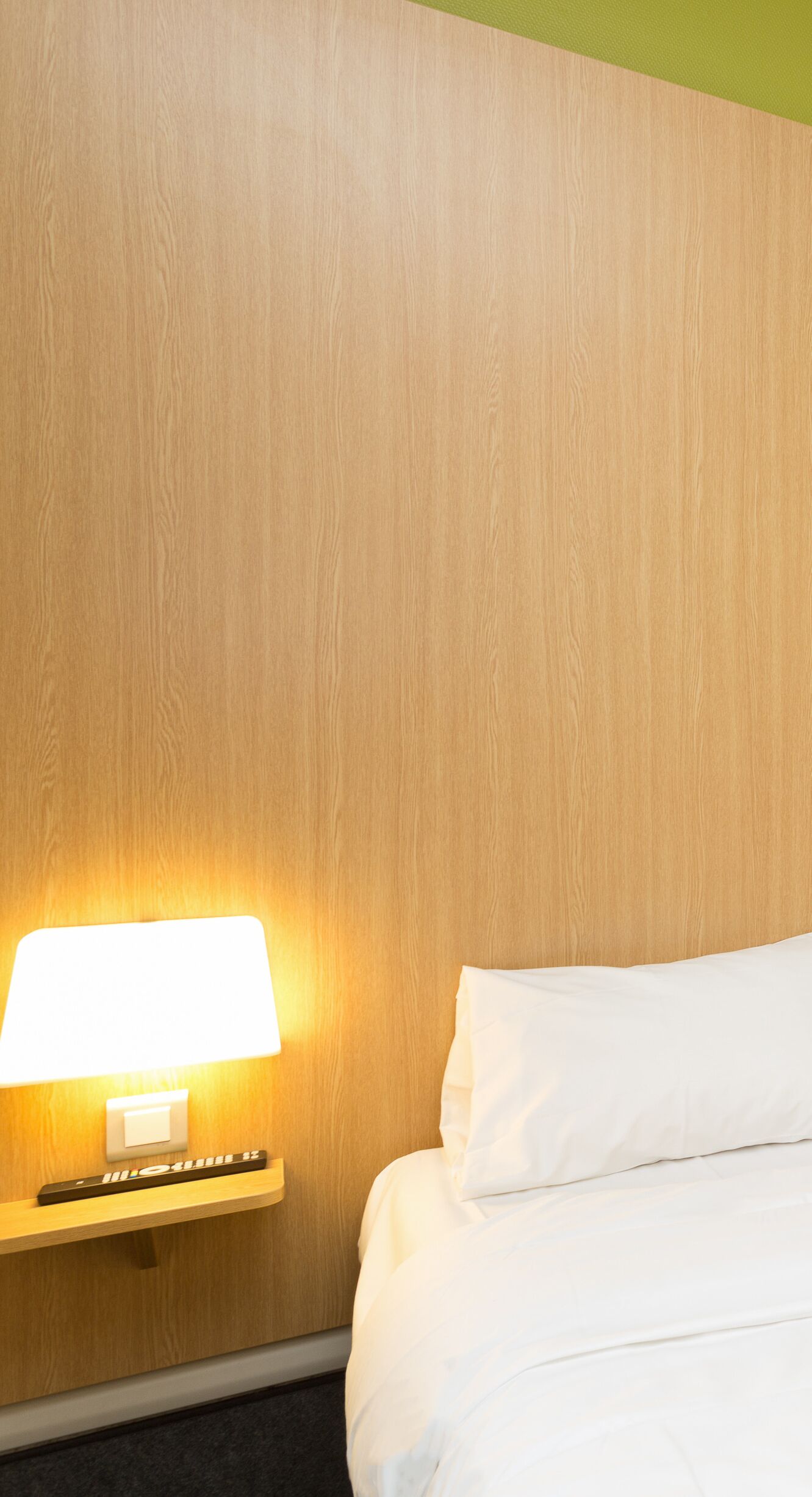 A hotel room with a wooden headboard, a bedside lamp, a remote control, and a neatly made bed.