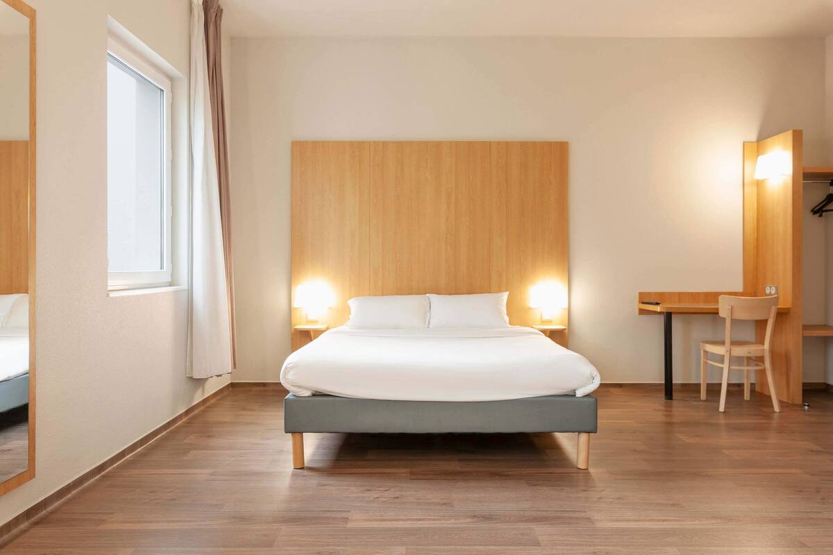Moderni hotellihuone, jossa on puinen sängynpääty, yksi sänky, työpöytä tuolilla ja suuri peili.