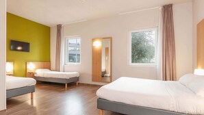 A modern hotel room with three beds, wooden flooring, large windows, and a green accent wall.