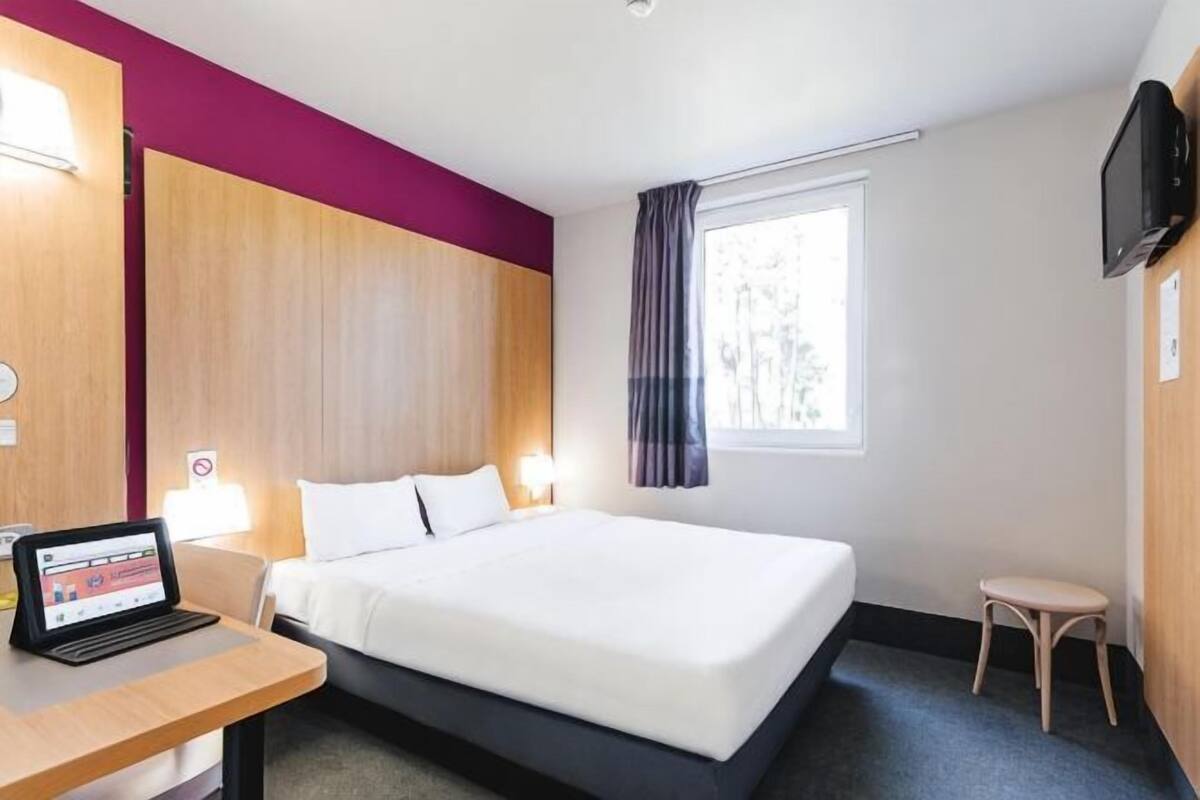 Hotellihuone, jossa on sänky, työpöytä kannettavalla tietokoneella, televisio ja ikkuna, jossa on verhot.