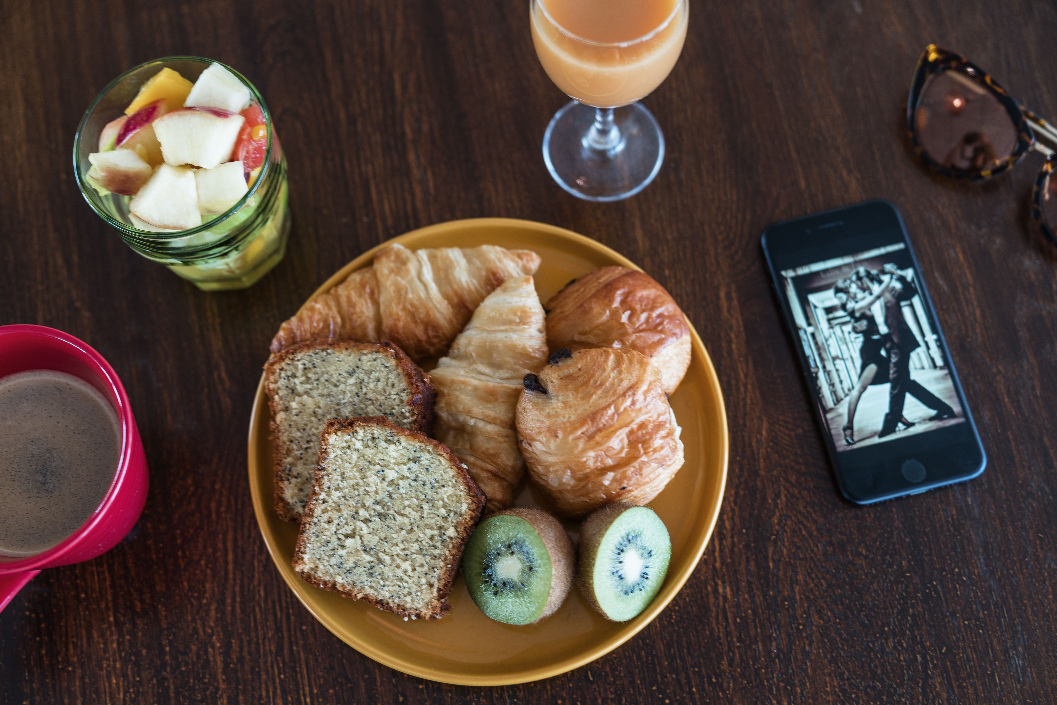 A plate with croissants, kiwi slices, and bread, a glass of juice, and a smartphone on a wooden table.