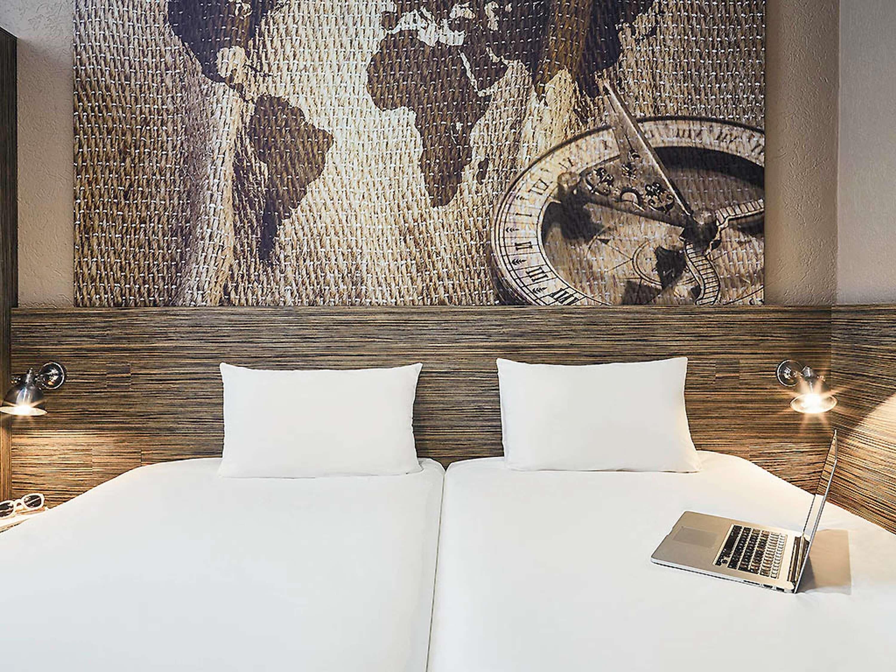 A hotel room with a large world map and clock on the wall, a bed with white linens, and a laptop on the bed.