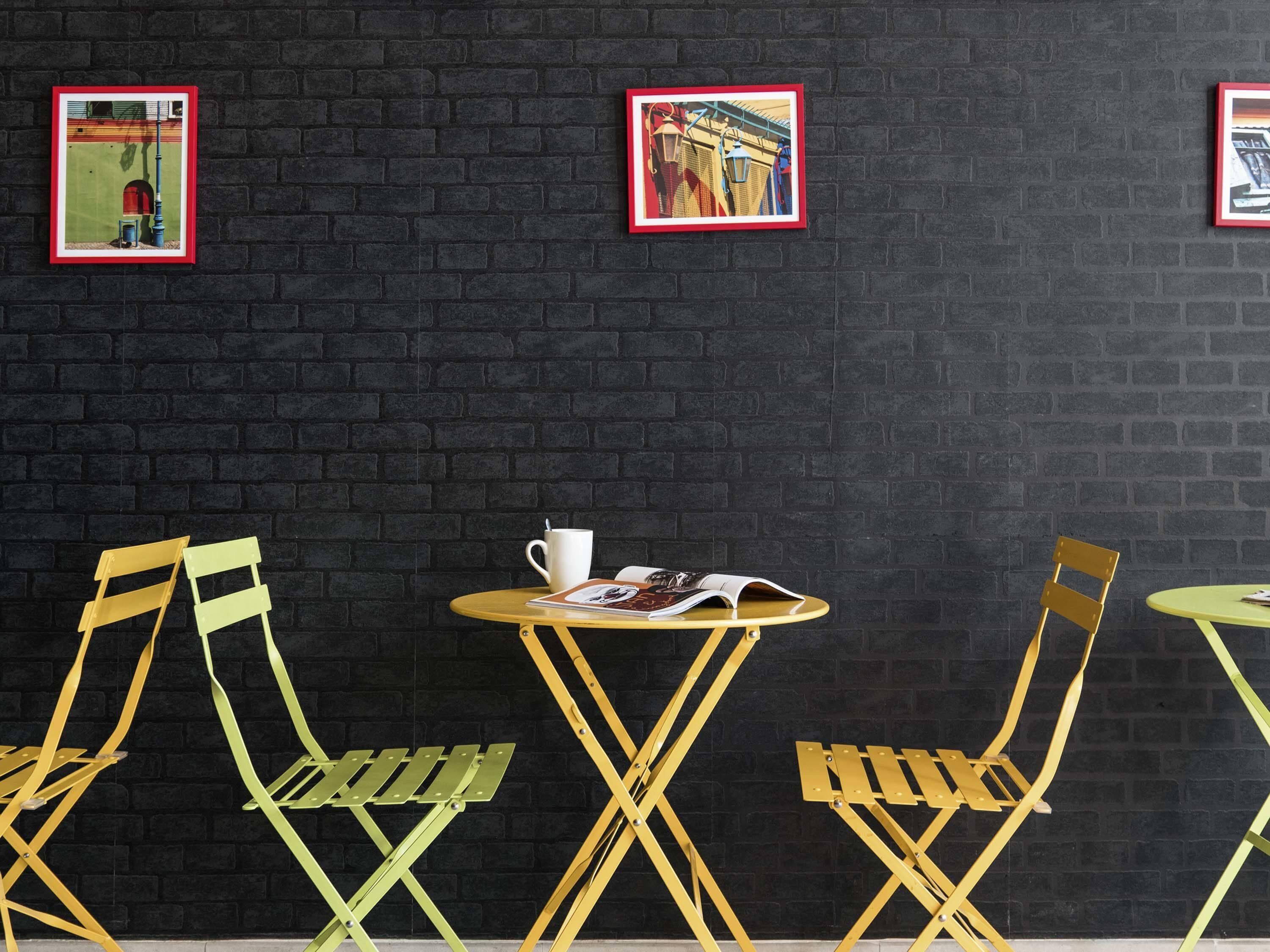 A café with black brick walls, yellow and green metal chairs, and two framed pictures.