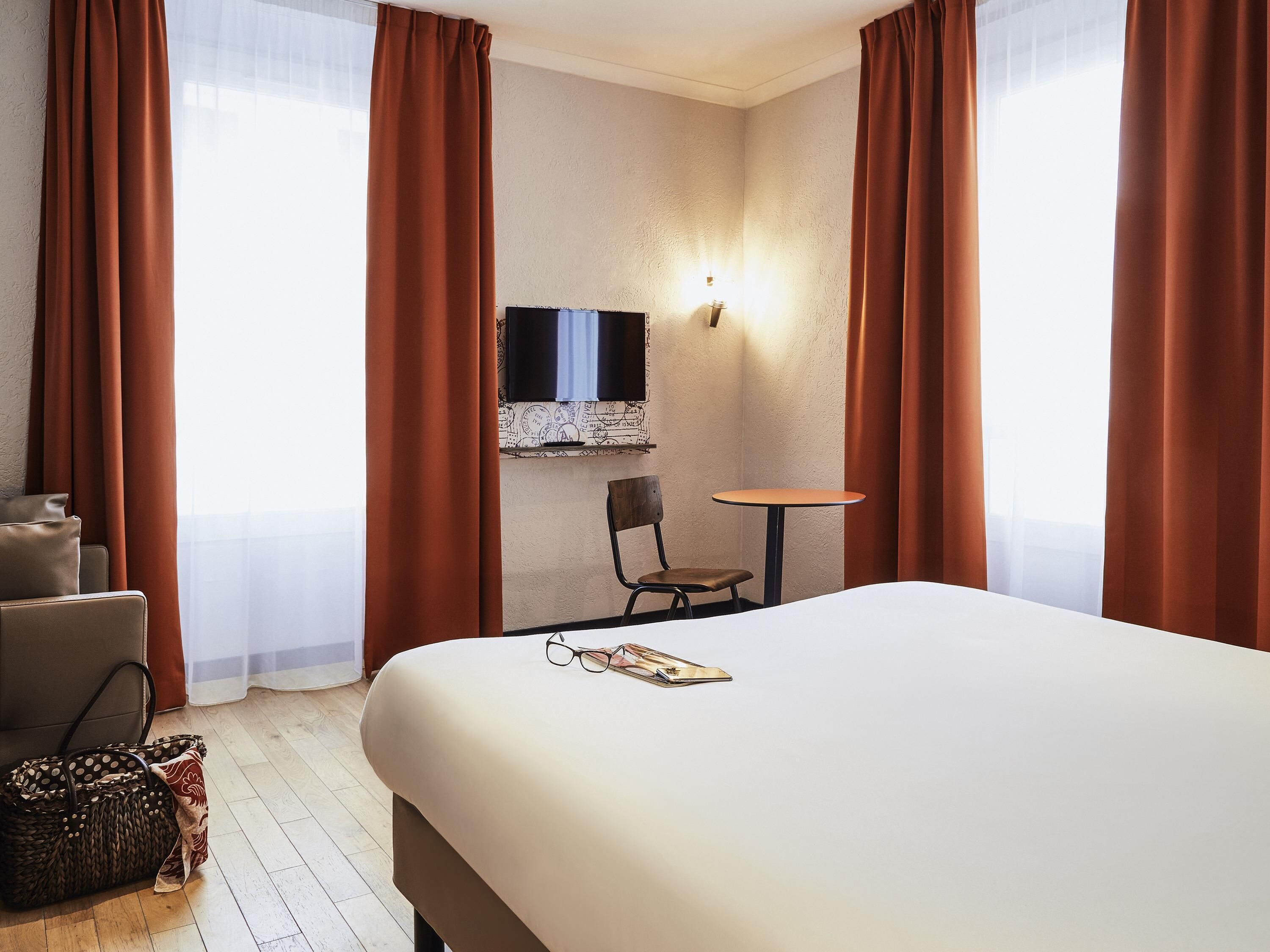 A hotel room with a bed, a chair, a small table, a TV mounted on the wall, and a window with curtains.