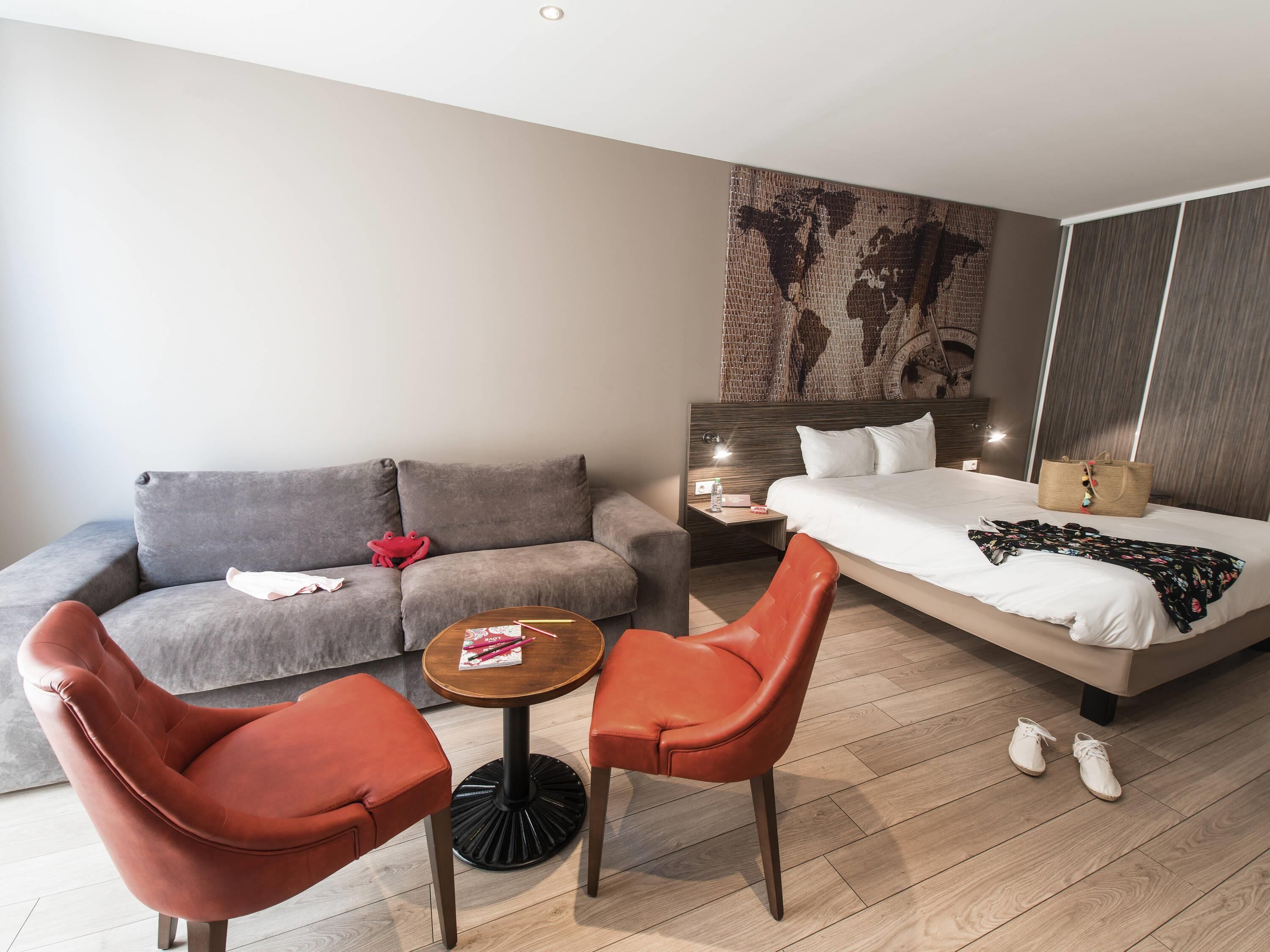 Een moderne hotelkamer met een grijze bank, twee rode stoelen, een klein rond tafeltje, een bed met een witte dekbedovertrek en een wereldkaart aan de muur.