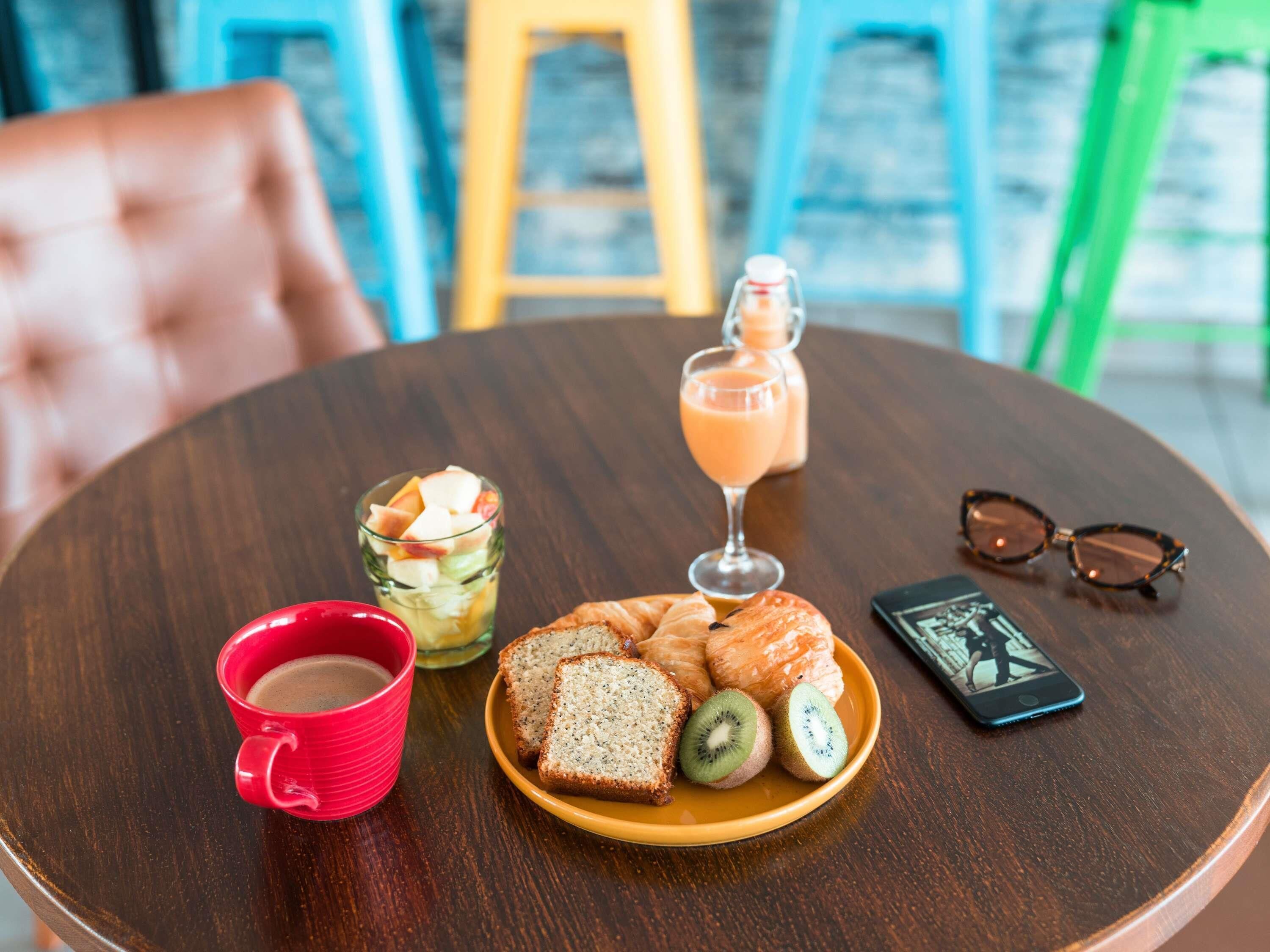 Una mesa de madera redonda con un plato de comida, un vaso de jugo, una taza roja y un teléfono inteligente.
