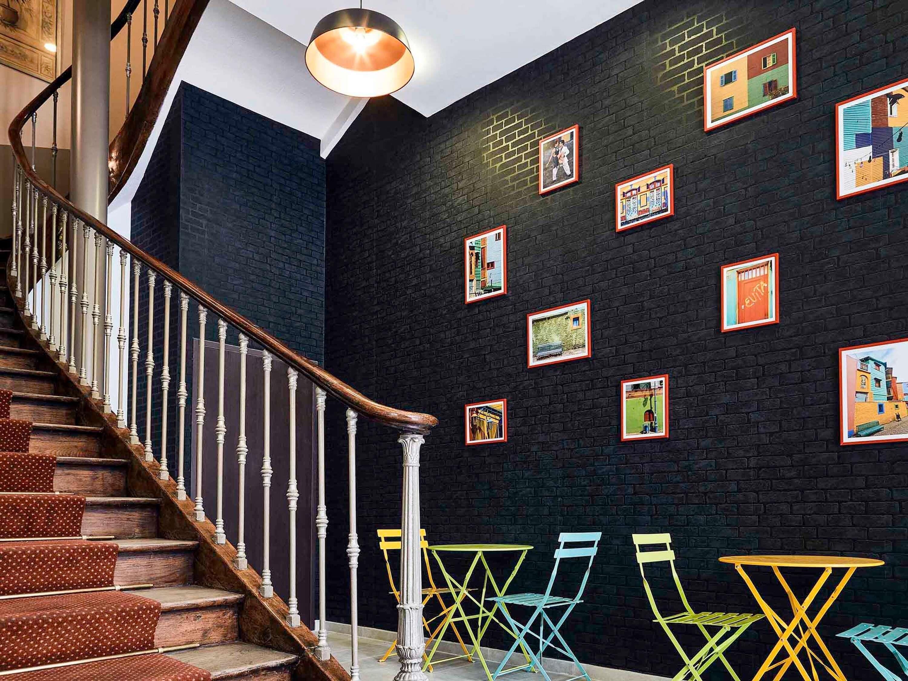 A staircase with wooden steps and white balusters, a black brick wall with framed pictures, and colorful chairs in the foreground.