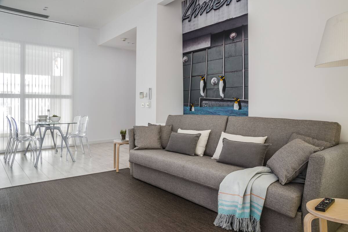 Un salon moderne avec un canapé gris, une table à manger en verre transparent et un tableau représentant des pingouins accroché au mur.