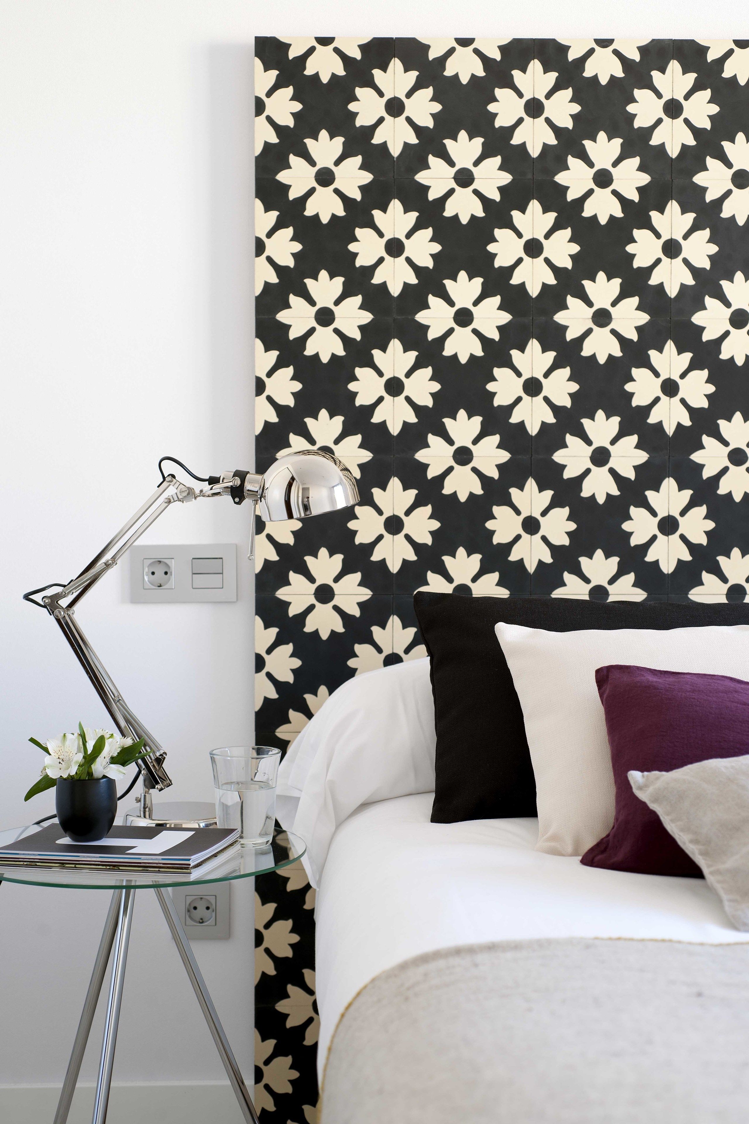 A neatly arranged bedroom with a patterned wallpaper, a bedside table with a lamp, and a bed with pillows.