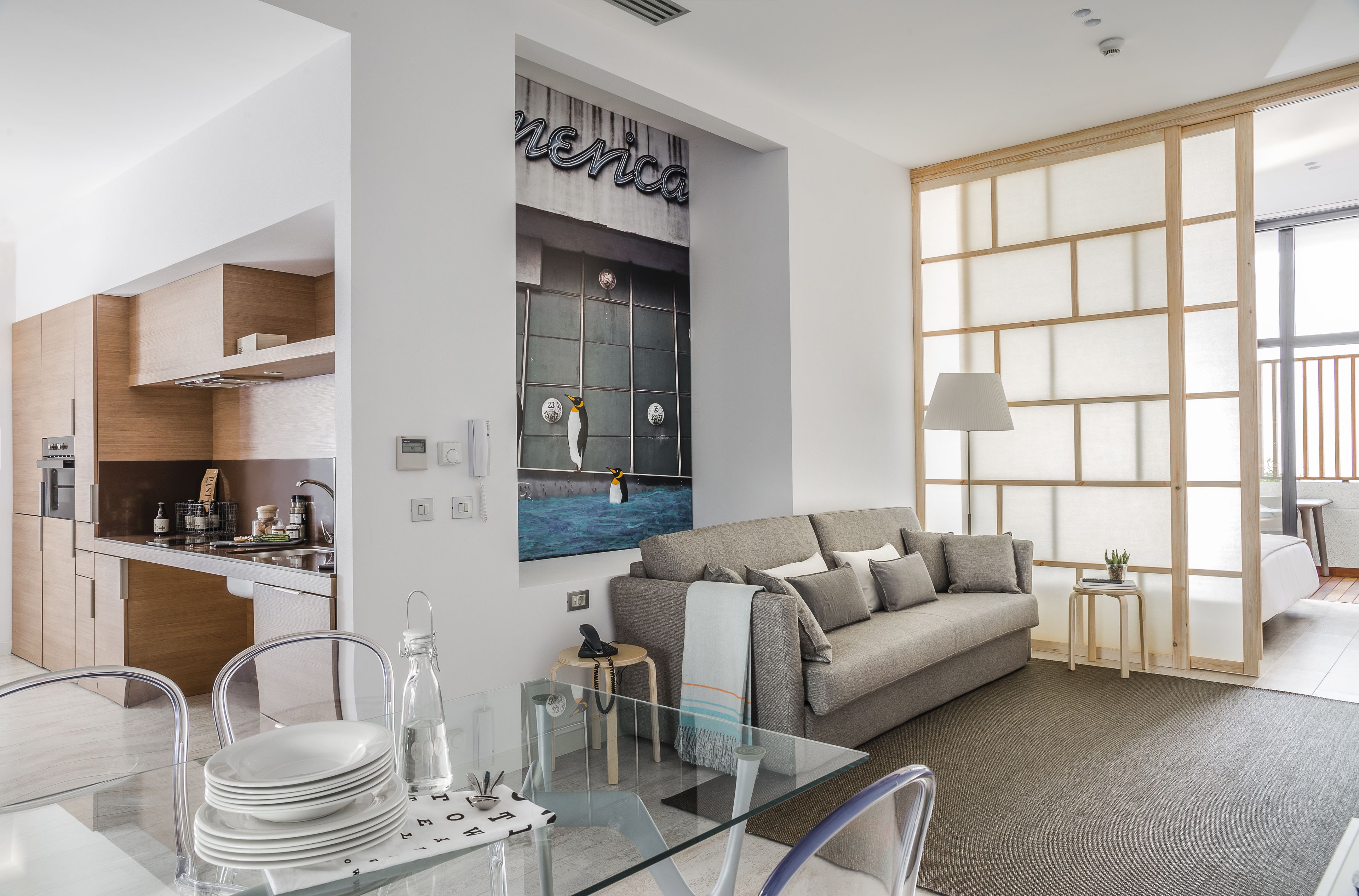 A modern living room with a sofa, a glass dining table, and a kitchen area with wooden cabinets.