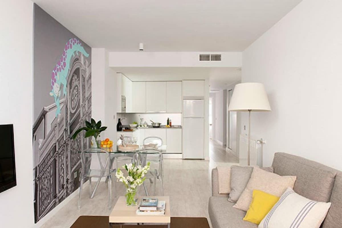 A modern living room with a sofa, coffee table, and clear dining table. A kitchen is visible in the background with white cabinets and a refrigerator.