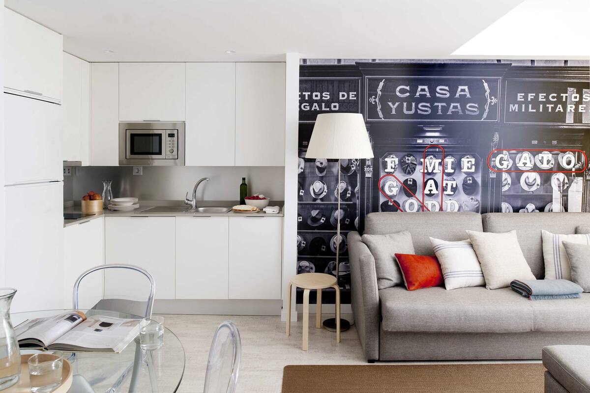 Une cuisine moderne avec des armoires blanches, un four à micro-ondes et un évier. On y trouve un coin repas avec une table en verre, des chaises et un magazine. Un canapé gris avec des coussins est placé contre le mur, et une lampe se trouve sur une table d’appoint.