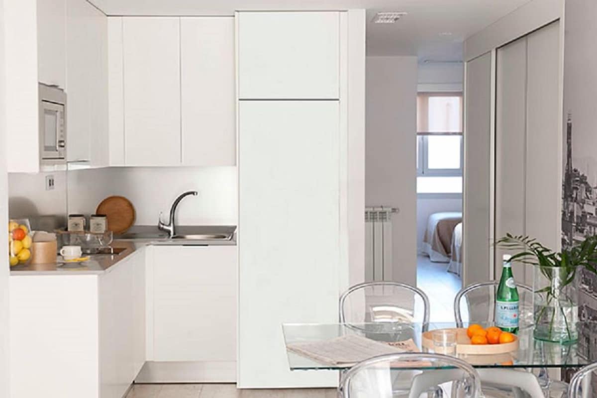 A compact kitchen with white cabinets, a sink, and a glass table with oranges and a bottle.