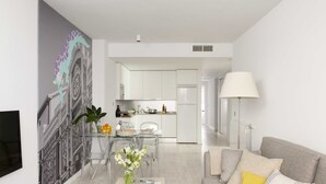 Una sala de estar moderna con un sofá gris, una mesa de centro con libros y flores, una mesa de comedor de vidrio transparente, y una cocina al fondo.