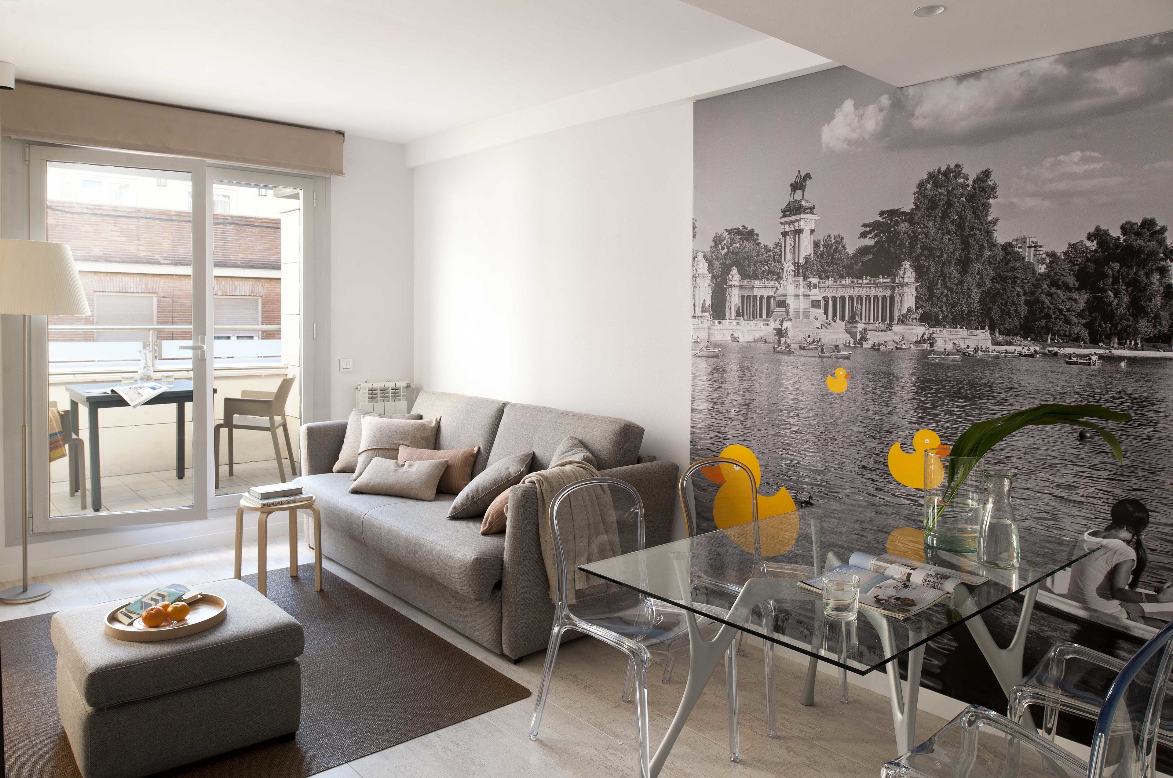 A modern living room with a grey sofa, clear glass dining table, and a large black-and-white mural of a historic building by a body of water.