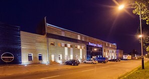 Un edificio con un cartel que dice "Boutique Hotel's" de noche, con un diseño moderno, ventanas grandes y un estacionamiento con varios autos.