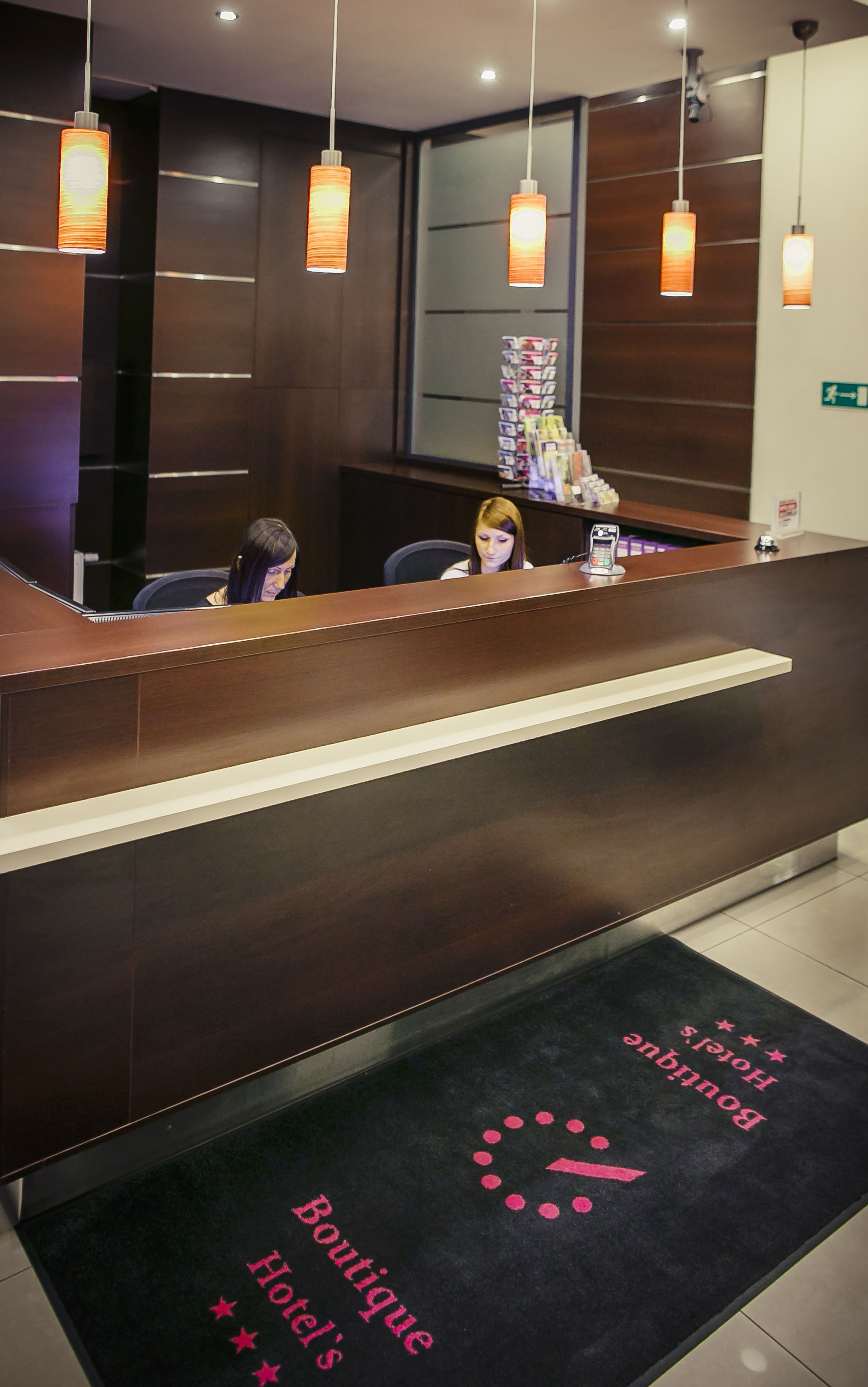 Zona de recepción de un hotel con un mostrador de madera oscura, una alfombra negra con el texto 'Boutique Hotel' y luces colgantes.