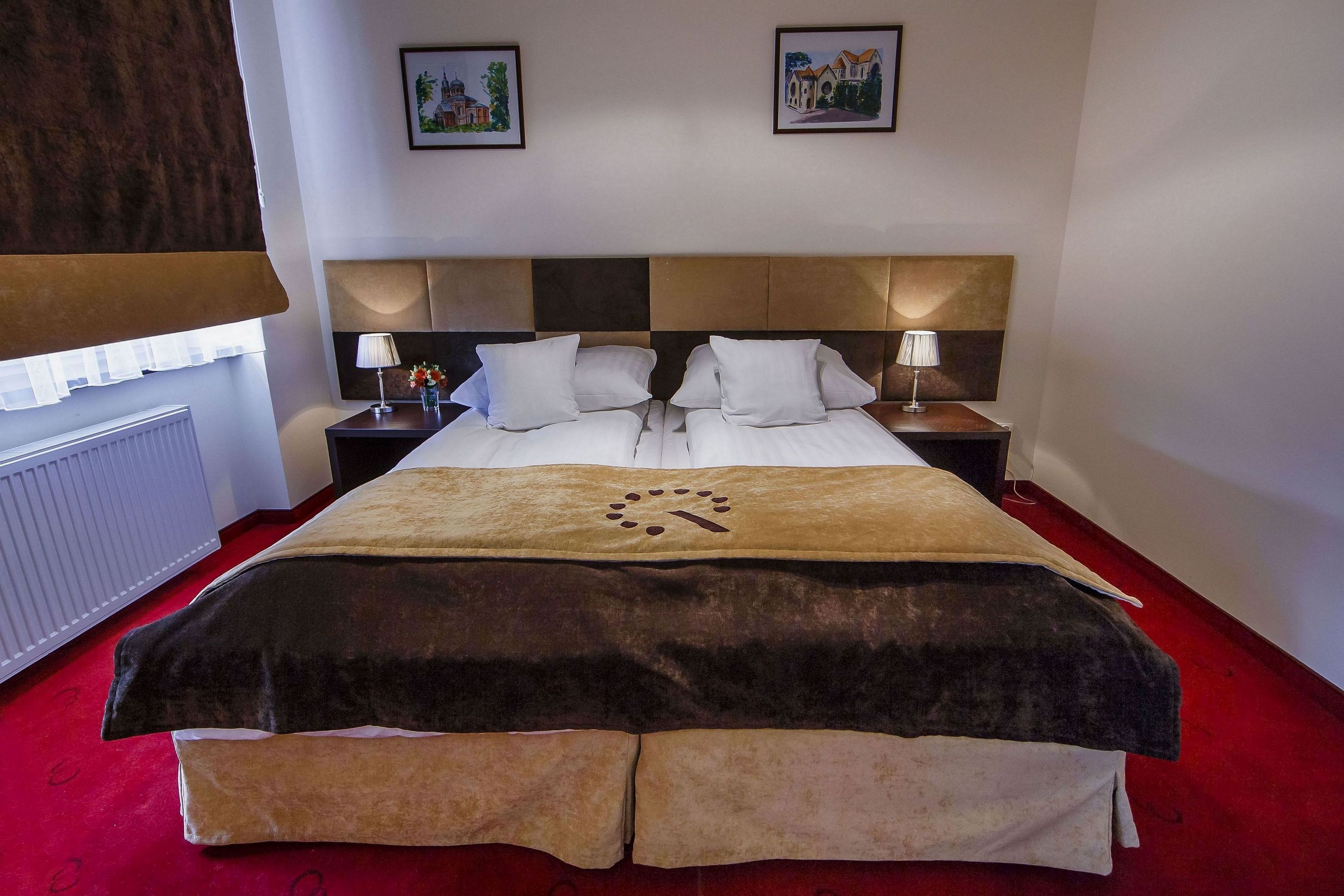 Habitación de hotel con una cama grande, dos mesitas de noche, una alfombra roja y dos cuadros en la pared.