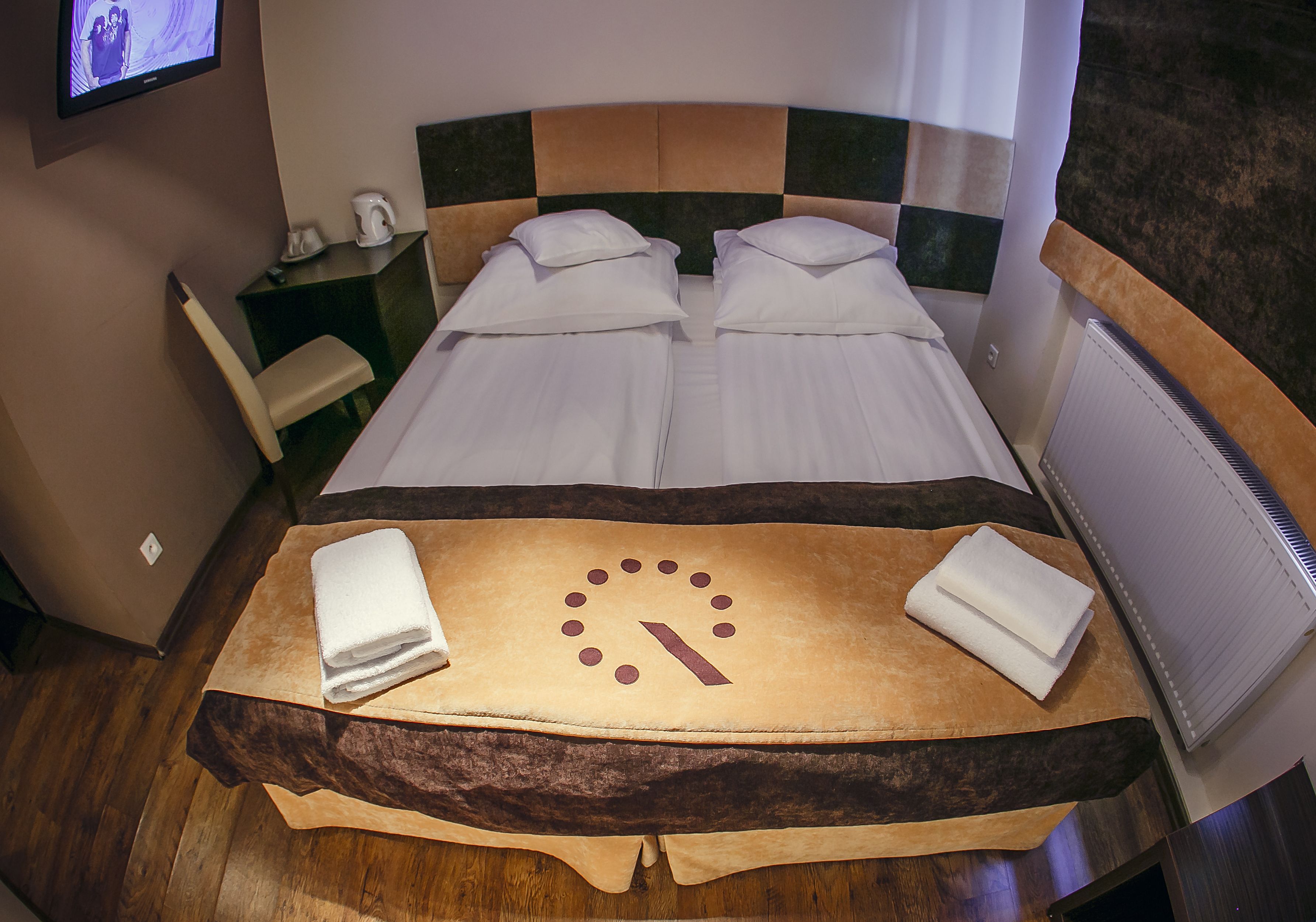 A double bed with a clock design on the bedspread, a wooden headboard, and a small table with towels.
