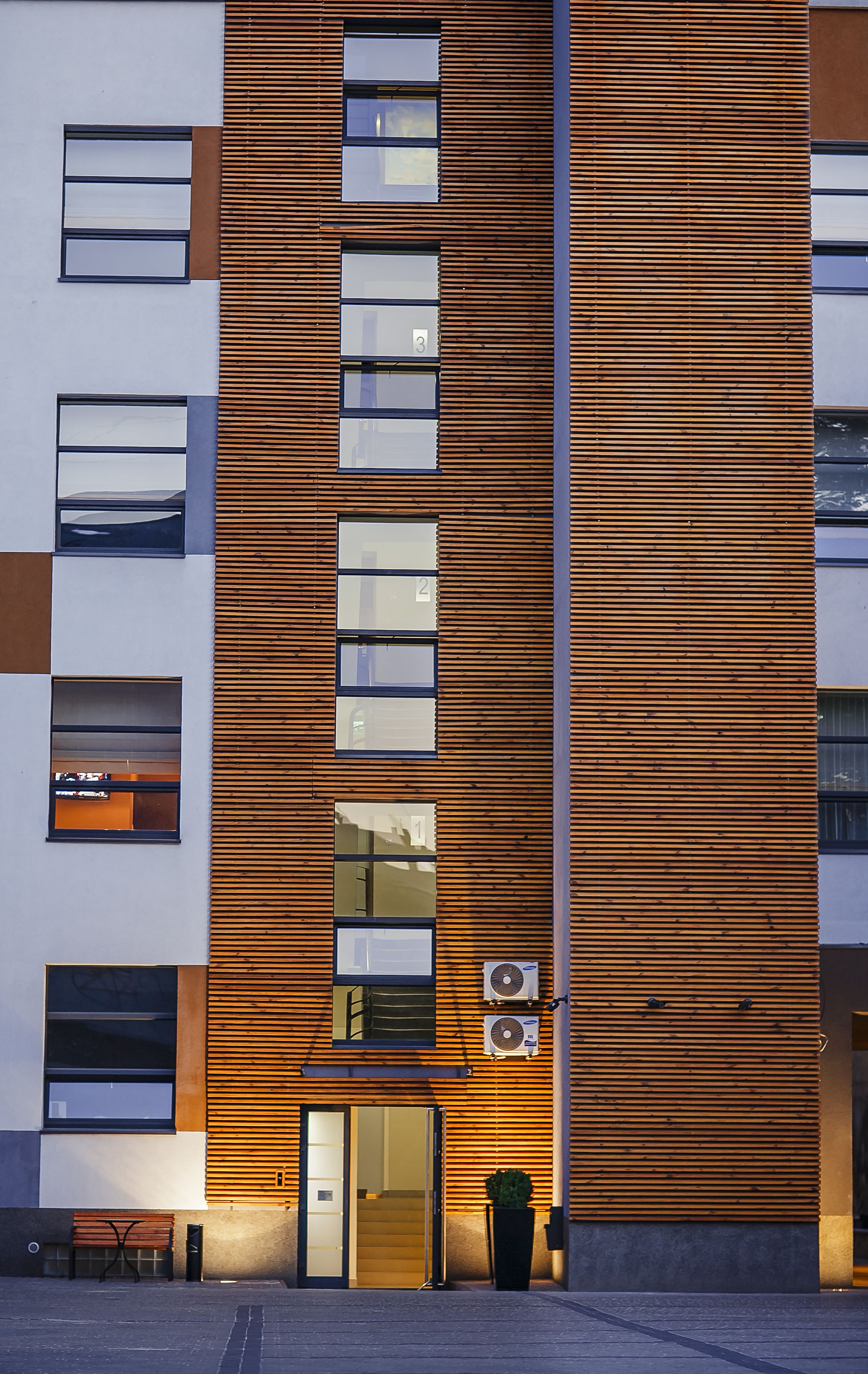 A modern building with a wooden facade, multiple windows, and an entrance with a bench and planter outside.