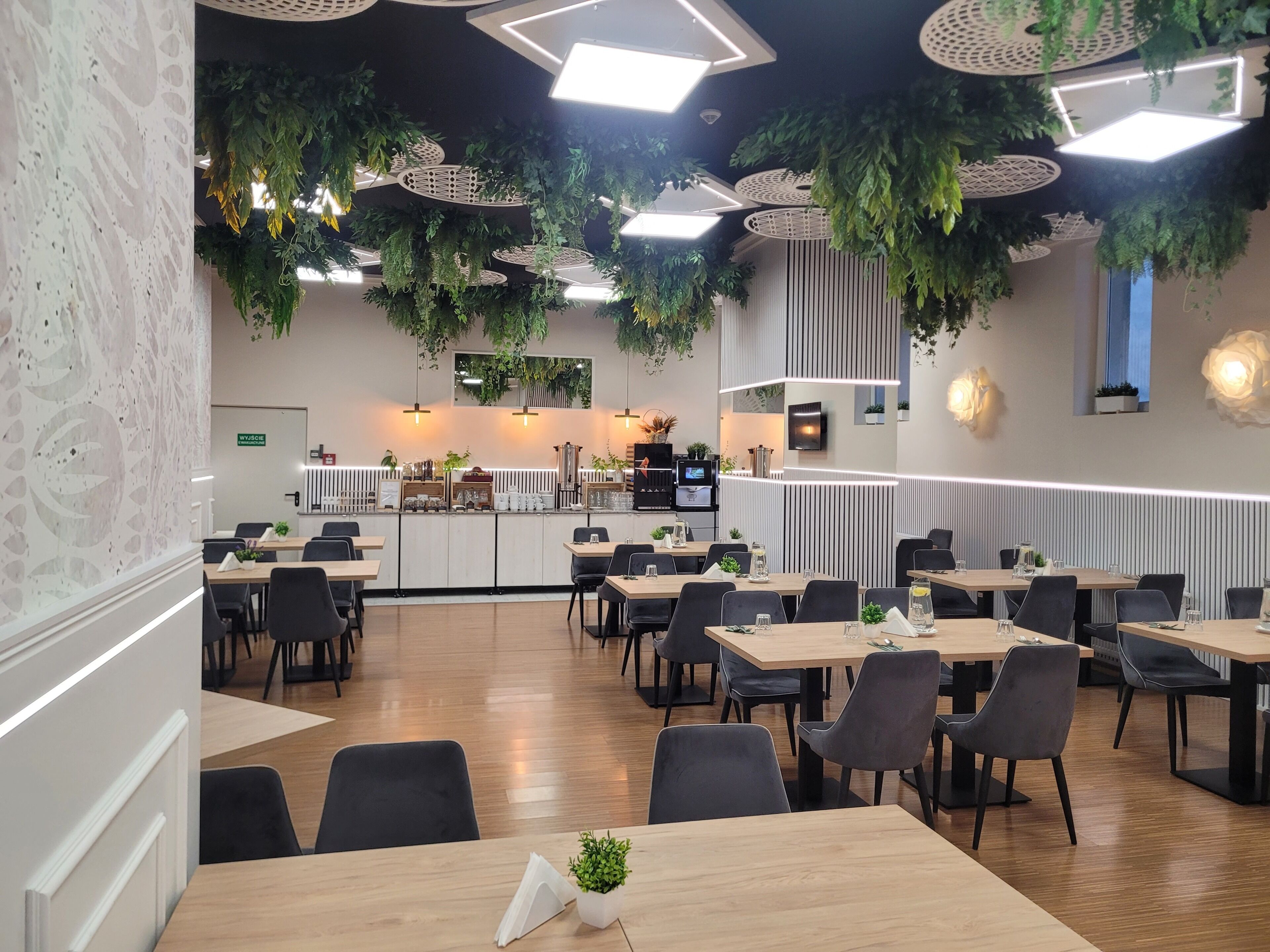 A modern restaurant interior with wooden tables and chairs, hanging plants, and a central counter area.