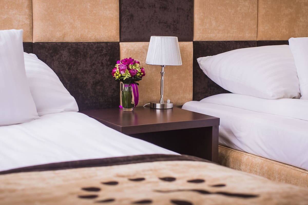 A hotel room with two beds, a nightstand with a lamp, and a vase with flowers.